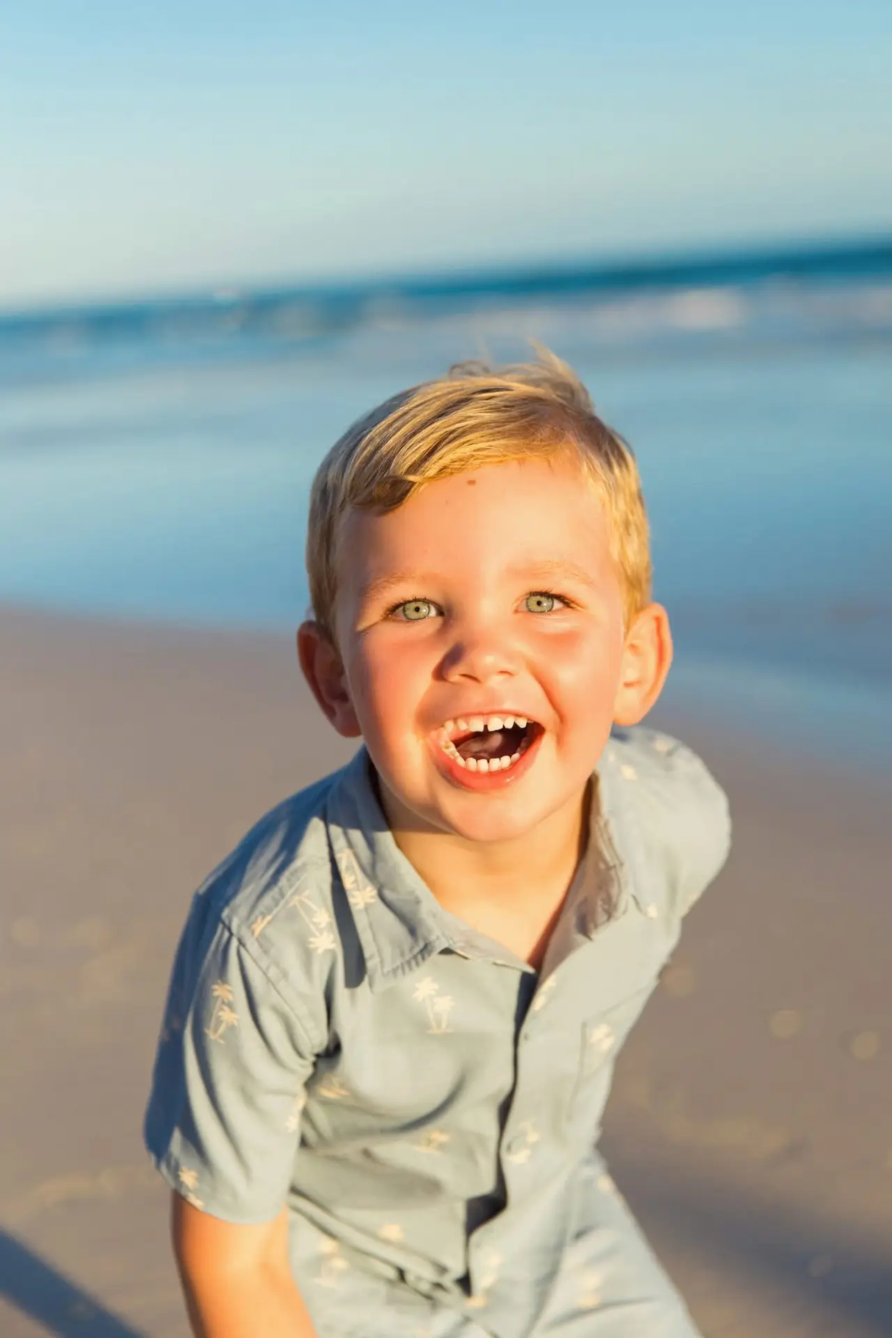 A young boy with blond hair and bright blue eyes grins widely at the camera on a sandy beach with the ocean in the background.