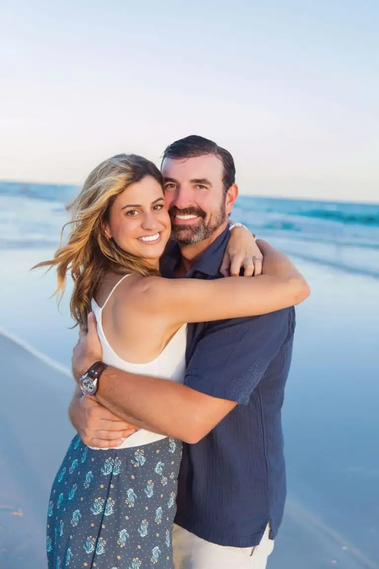A smiling couple embraces on a beach with the ocean waves in the background under a soft sky.