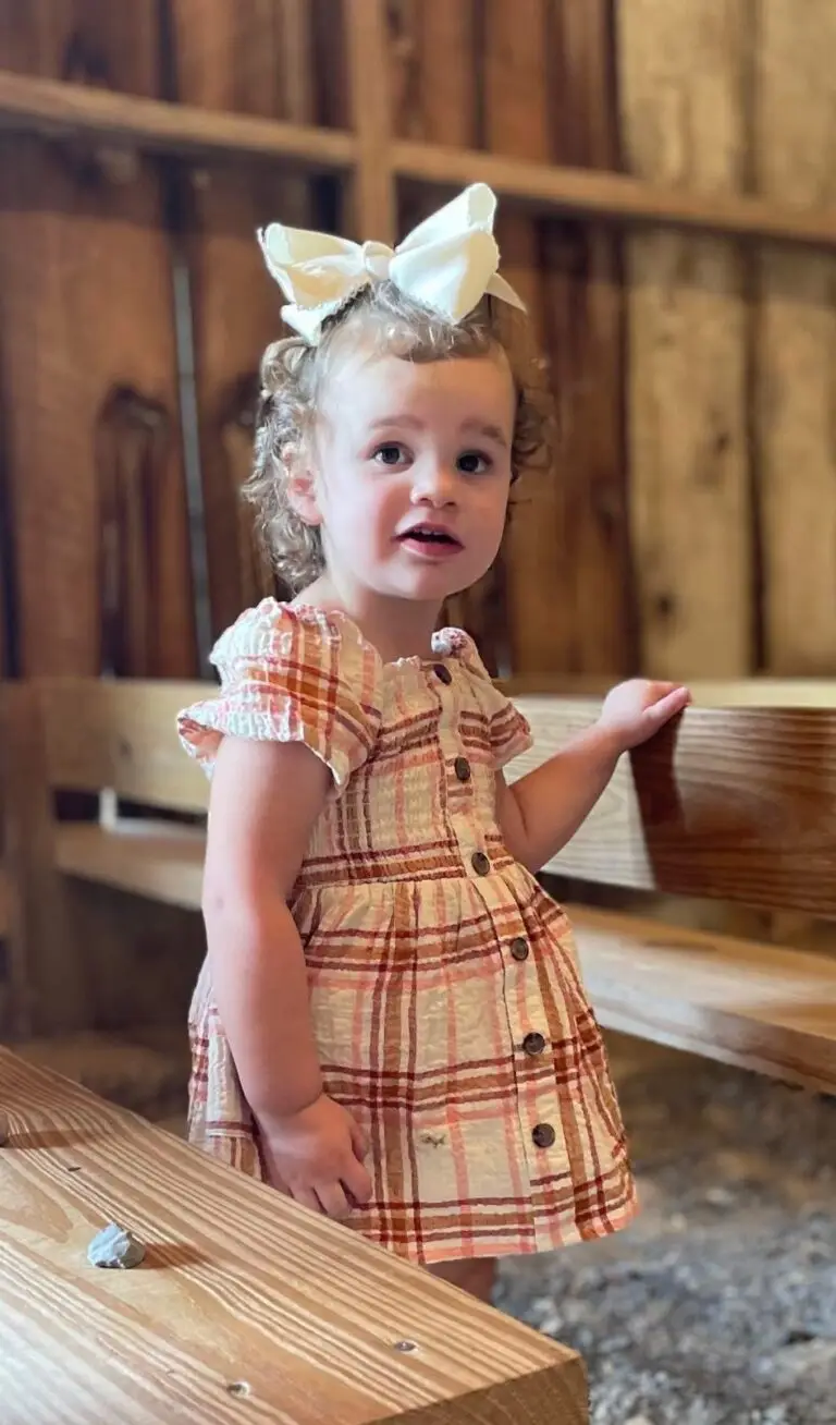 A young child with curly hair and a large white bow in a plaid dress stands next to a wooden bench.