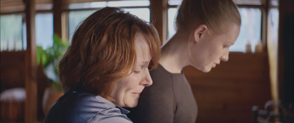 A close-up side profile of a middle-aged woman with auburn hair resting her head gently against the shoulder of a younger woman with blonde hair, both with serene or introspective expressions. They appear to be in a warm, possibly wooden interior space.