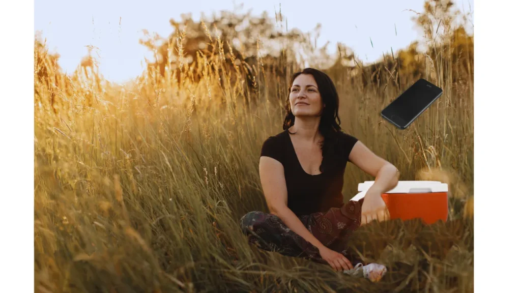 A woman with dark hair, wearing a dark t-shirt and patterned pants, sits cross-legged in a sun-drenched field of tall golden grass. She is looking up and to her right with a calm, content expression, bathed in the warm glow of what appears to be late afternoon or sunset.