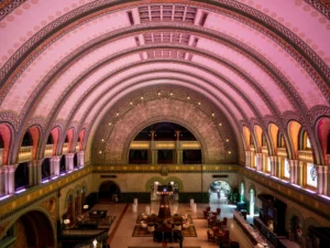 The expansive, arched ceiling of Union Station's grand hall glows with pink and purple light, illuminating intricate architectural details, while a large, ornate window overlooks the bustling lower level.
