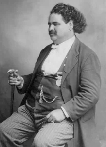 A black and white portrait of John William "Blind" Boone, a large man with dark curly hair, a mustache, and a slight smile, wearing a suit with a bow tie and a medal, holding a cane in his right hand.