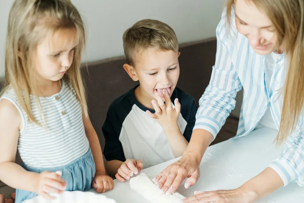 A woman and two young children are focused on rolling dough on a white table, with one child licking their fingers.