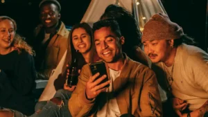 A diverse group of five young adults smiles and laughs while looking at a smartphone, gathered outdoors around a tent with string lights, suggesting a social evening or camping trip.