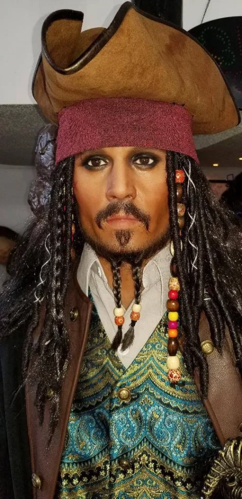 A highly detailed wax figure of Captain Jack Sparrow, recognizable by his tricorn hat, dreadlocks with beads, smudged eyeliner, mustache, and goatee, wearing a turquoise and gold patterned vest.