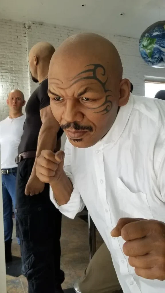 A close-up of a highly realistic wax figure of Mike Tyson, with his signature facial tattoo, scowling expression, and fists clenched in a boxing stance, wearing a white button-up shirt. Other wax figures are blurred in the background.