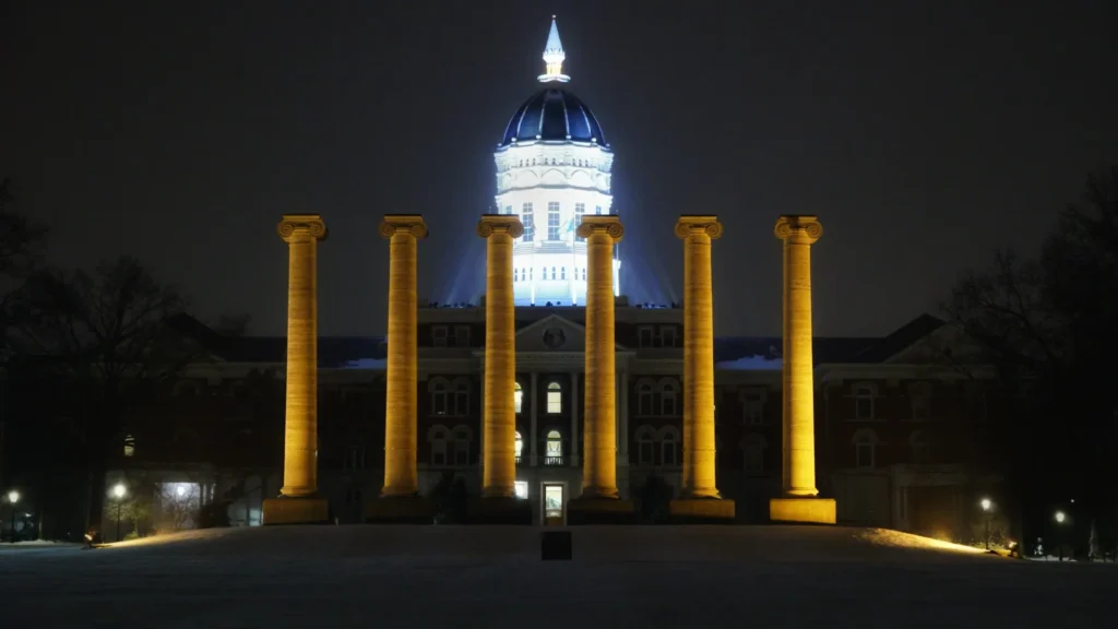 Six illuminated, golden-toned neoclassical columns stand in the foreground against a dark night sky, with the brightly lit dome and facade of Jesse Hall, a large university building, visible behind them.