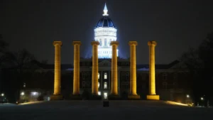 Six illuminated, golden-toned neoclassical columns stand in the foreground against a dark night sky, with the brightly lit dome and facade of Jesse Hall, a large university building, visible behind them.