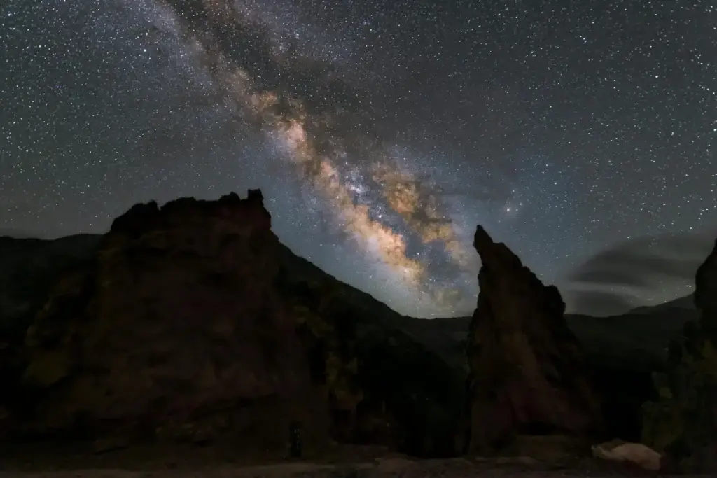The vibrant band of the Milky Way galaxy arches across a star-filled night sky above dark, rugged rock formations.