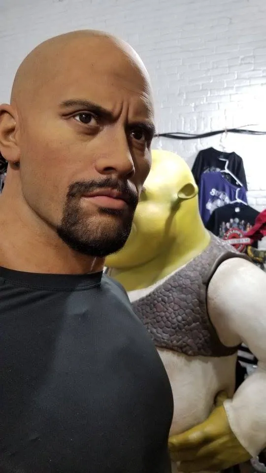 A realistic wax figure of Dwayne "The Rock" Johnson, bald and with a goatee, wearing a black shirt and looking intensely to the side. Behind him, part of a Shrek character costume or figure is visible.